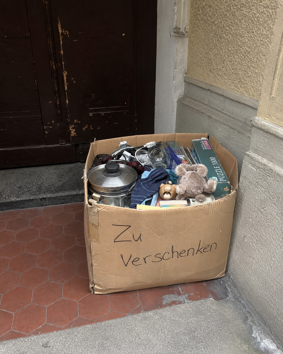 Gepackte Zu-verschenken-Kiste mit Topf, Gläsern, Spielzeug, Kleidung und Puzzle vor einem Hauseingang.